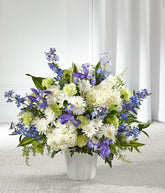 A standing basket of white hydrangeas, blue delphinium, iris, white poms, green carnations, and fresh floral greens in a white plastic urn