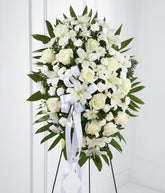 A standing spray made up of white lilies, roses, mums, and carnations, with fresh floral greens and a white ribbon