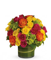 A floral arrangement featuring green hydrangea, hot pink roses, yellow roses, and bi-color roses in a clear glass vase.
