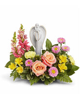 Arrangement featuring peach roses, yellow alstroemeria, yellow carnations, hot pink snapdragons, pink asters, and floral greenery with a ceramic figurine atop the arrangement