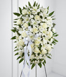 A standing spray made up of white lilies, roses, mums, and carnations, with fresh floral greens and a white ribbon