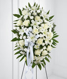A standing spray made up of white lilies, roses, mums, and carnations, with fresh floral greens and a white ribbon