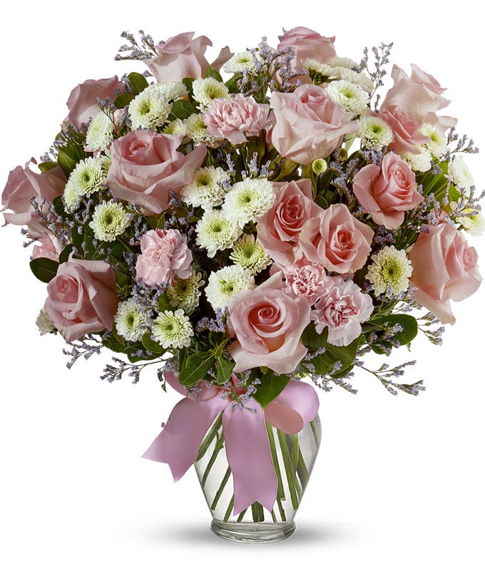 Floral arrangement featuring pink roses, white roses, mini carnations, and green poms in a clear glass vase with a bow.