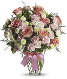 Floral arrangement featuring pink roses, white roses, mini carnations, and green poms in a clear glass vase with a bow.