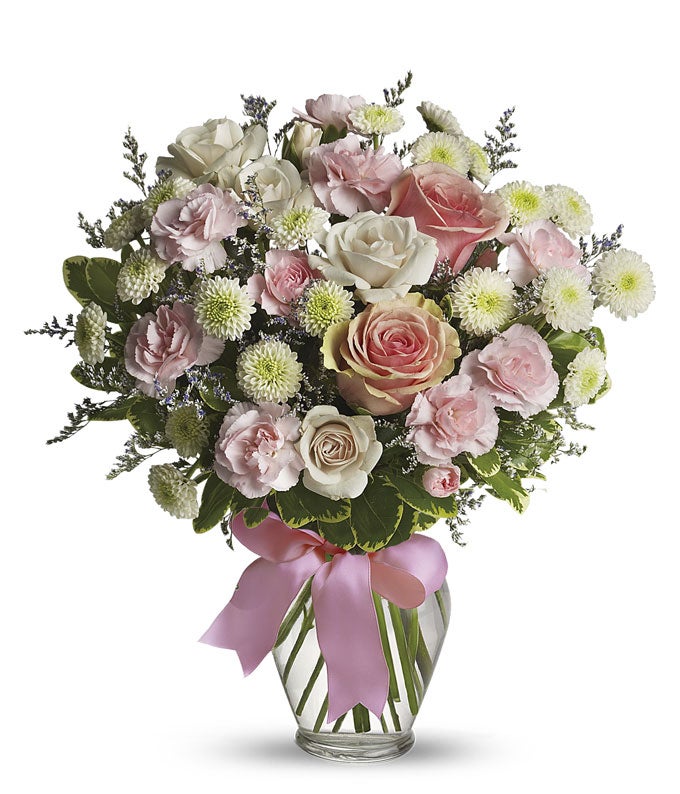 Floral arrangement featuring pink roses, white roses, mini carnations, and green poms in a clear glass vase with a bow.
