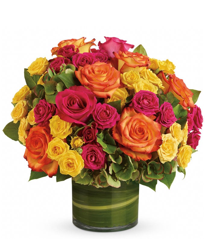 A floral arrangement featuring green hydrangea, hot pink roses, yellow roses, and bi-color roses in a clear glass vase.