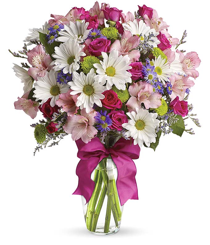 A cheerful floral arrangement featuring pink spray roses, white daisies, green button poms, purple Monte Casino, and limonium, all beautifully arranged in a glass vase with a hot pink ribbon. This vibrant mix offers a playful and lively display.