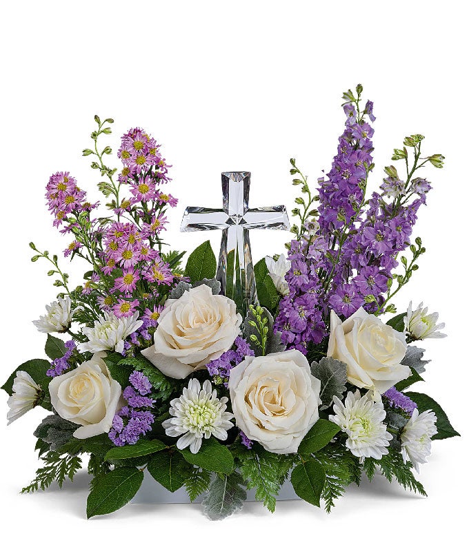 A graceful floral arrangement showcasing crème roses, lavender larkspur, lavender asters, lavender statice, white mums, and floral greens, elegantly accented with a crystal cross. This bouquet blends soft, calming colors for a serene display.