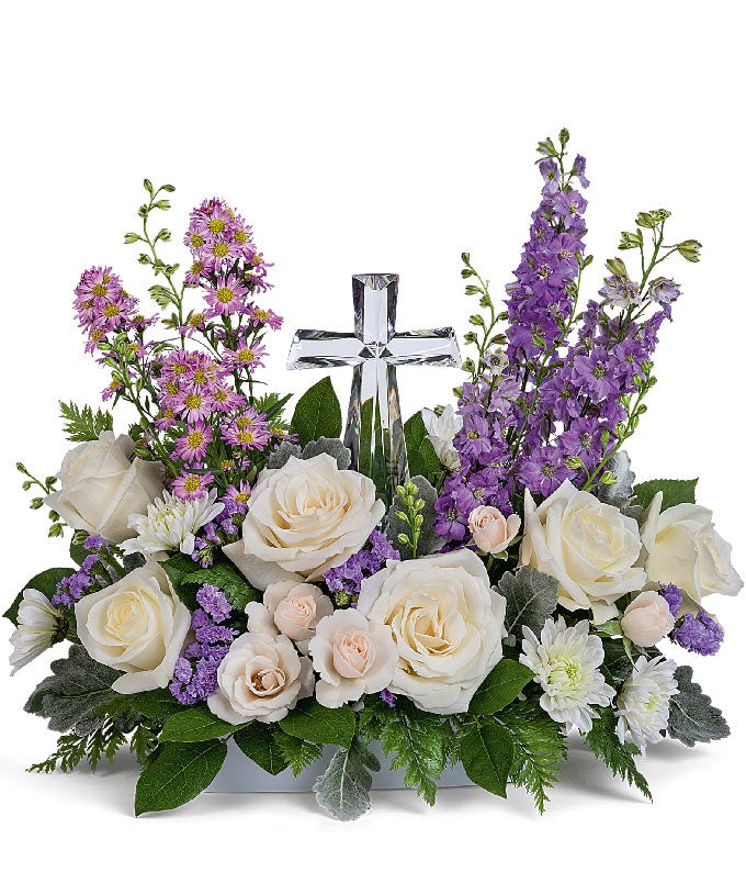 A graceful floral arrangement showcasing crème roses, lavender larkspur, lavender asters, lavender statice, white mums, and floral greens, elegantly accented with a crystal cross. This bouquet blends soft, calming colors for a serene display.