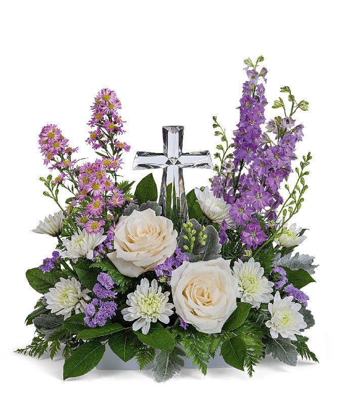 A graceful floral arrangement showcasing crème roses, lavender larkspur, lavender asters, lavender statice, white mums, and floral greens, elegantly accented with a crystal cross. This bouquet blends soft, calming colors for a serene display.