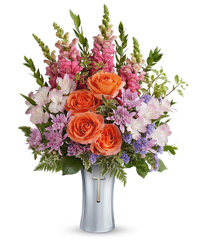 Orange roses, pink snapdragons, pink alstroemeria, lavender cushion spray chrysanthemums, purple statice, and assorted greenery arranged in a keepsake silver cross vase.