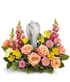 Arrangement featuring peach roses, yellow alstroemeria, yellow carnations, hot pink snapdragons, pink asters, and floral greenery with a ceramic figurine atop the arrangement 